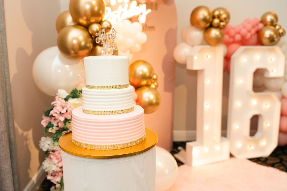 Elegant three-tier pink and white birthday cake with gold accents beside marquee number 16 balloons.