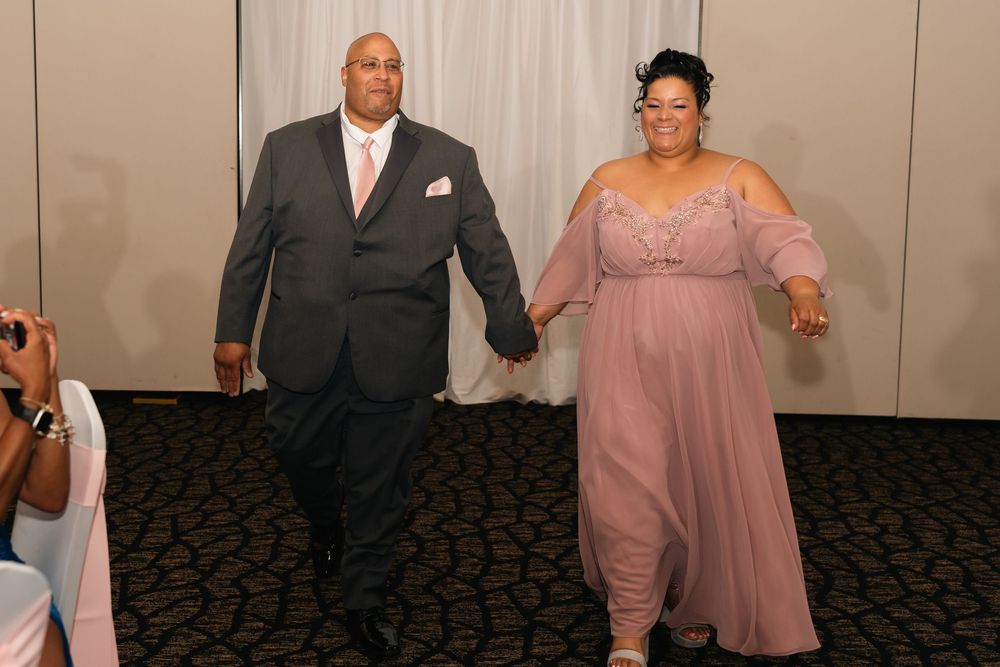 Couple in formal attire holding hands at a wedding reception, man in gray suit, woman in pink gown.