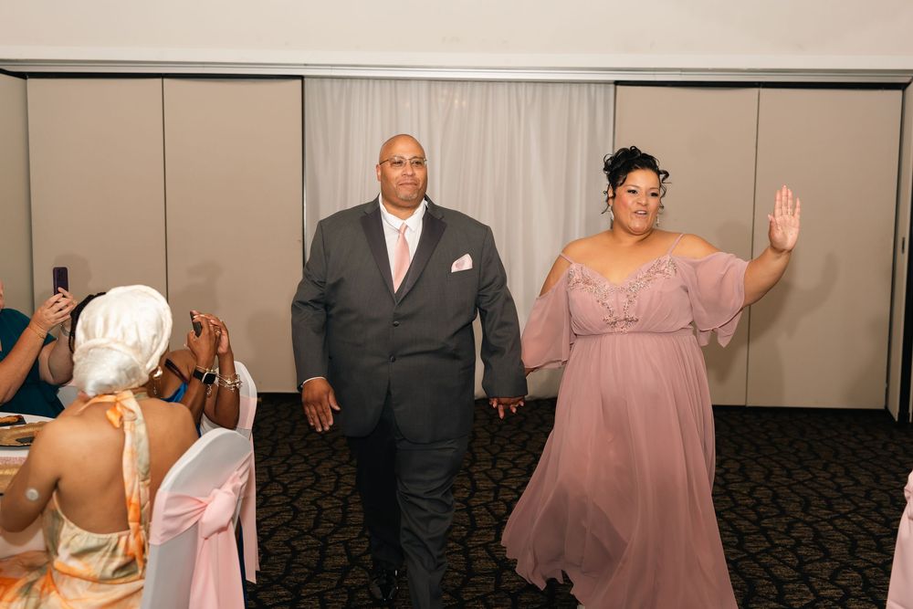 Couple making a grand entrance at a formal event, man in gray suit and woman in lavender gown waving.