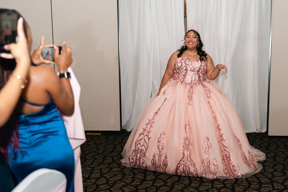 Young woman in pink ballgown quinceañera dress poses while guest photographs her at elegant celebration.