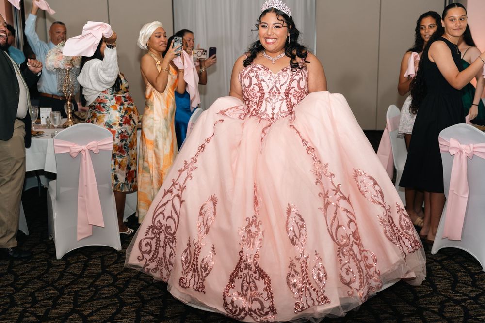 Young woman in pink quinceañera ballgown twirling at celebration party with guests cheering.