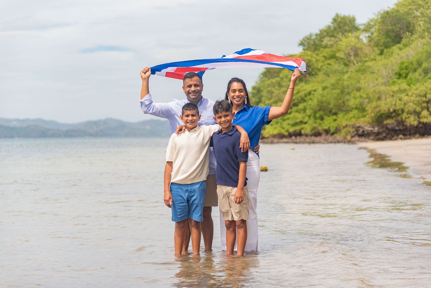 Family of four standing in ocean water holding a colorful flag high on a tropical beach.