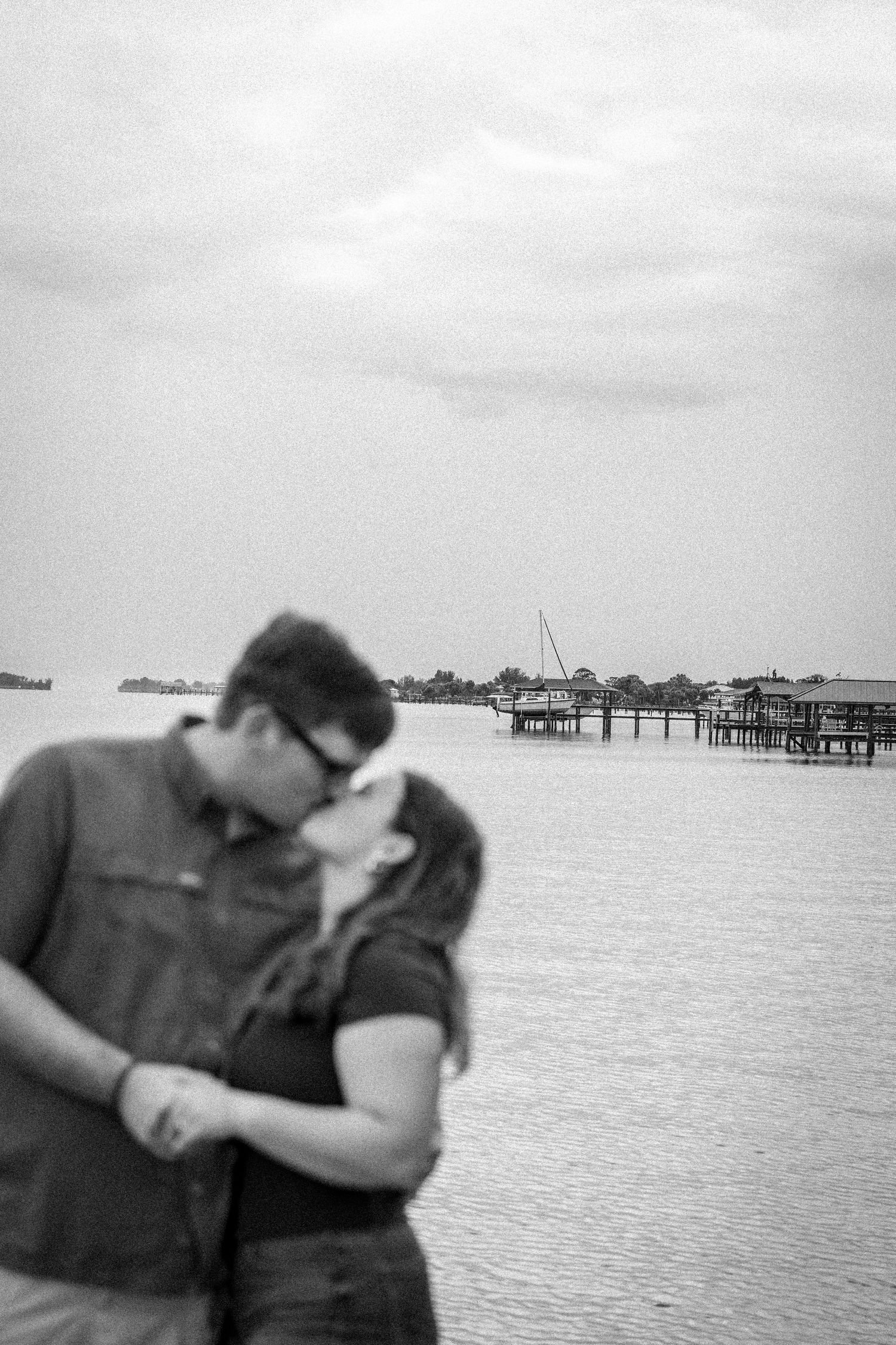 A couple shares a romantic kiss in a blurred black and white photo with a waterfront dock scene behind them.