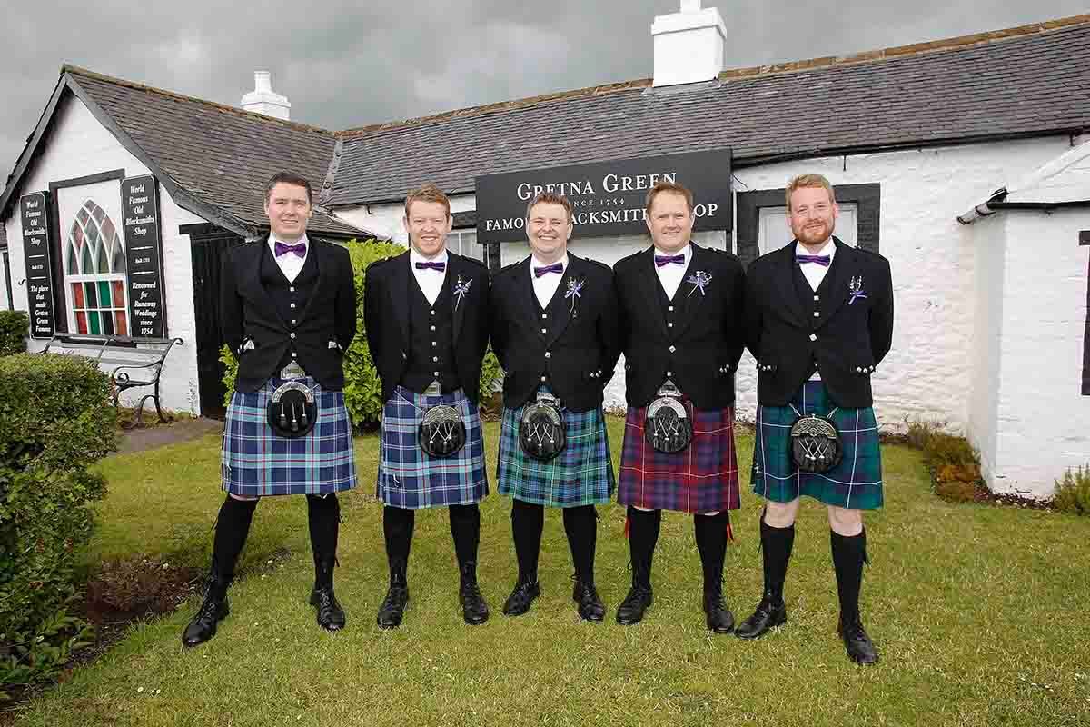 Five men in traditional Scottish kilts posing outside Gretna Green Famous Blacksmiths Shop on a cloudy day.
