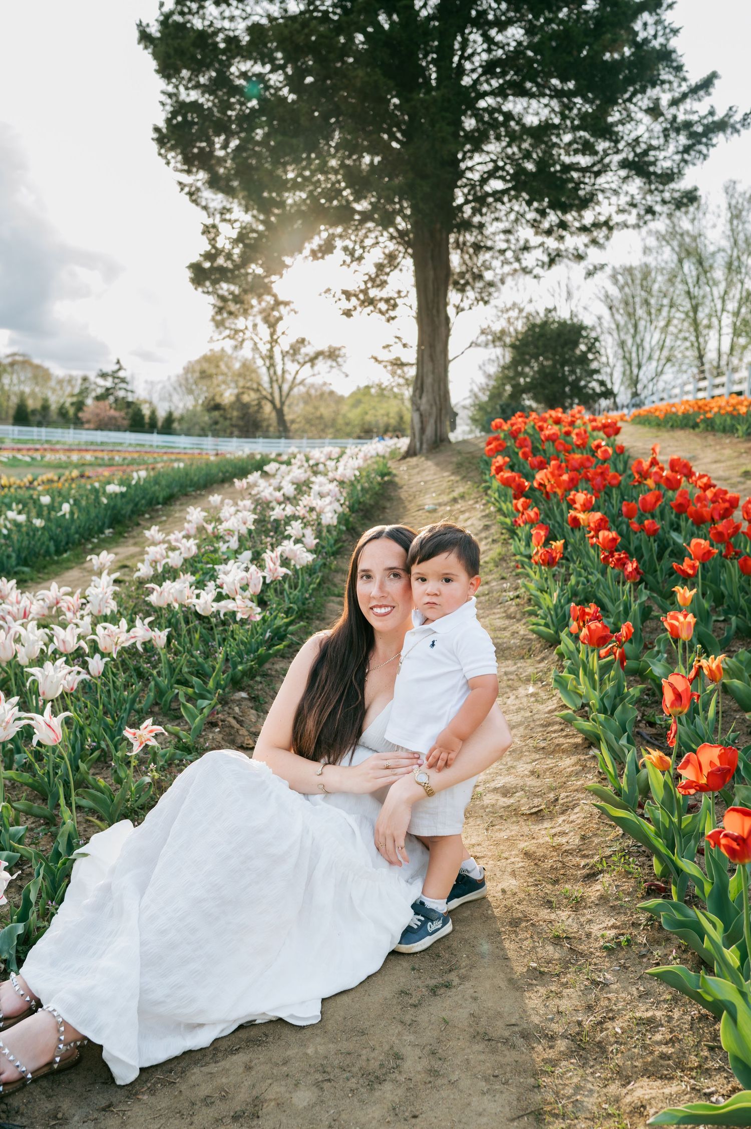 Mother in white dress sits with toddler son among vibrant tulip rows in sunny garden Greensboro NC