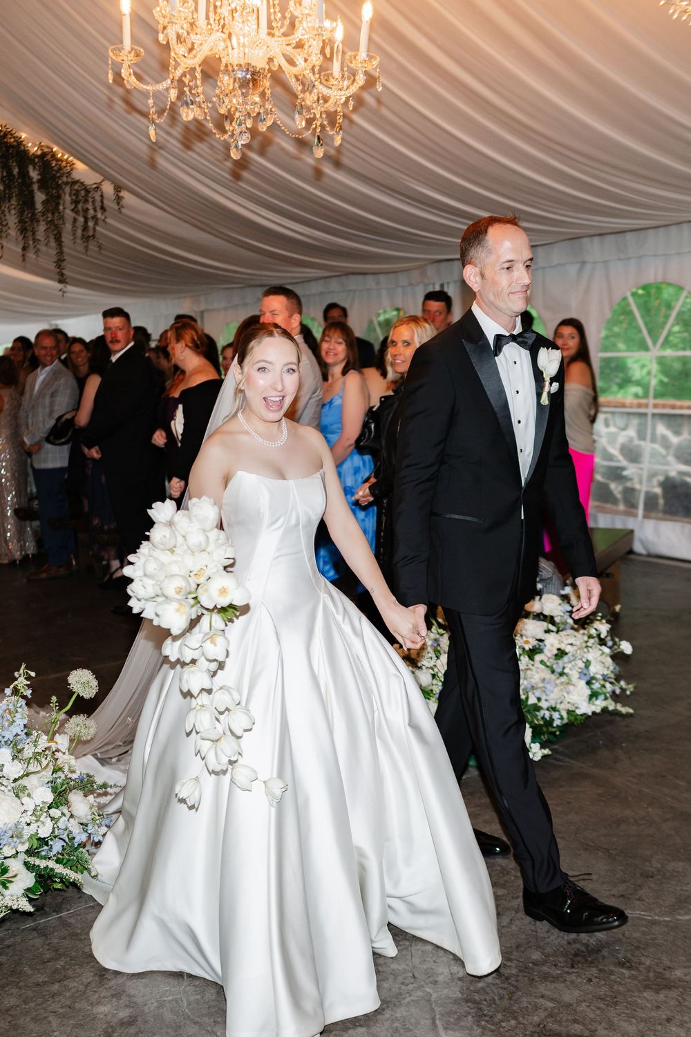 Joyful bride in white ballgown with cascading bouquet walks with groom after wedding ceremony in tent.