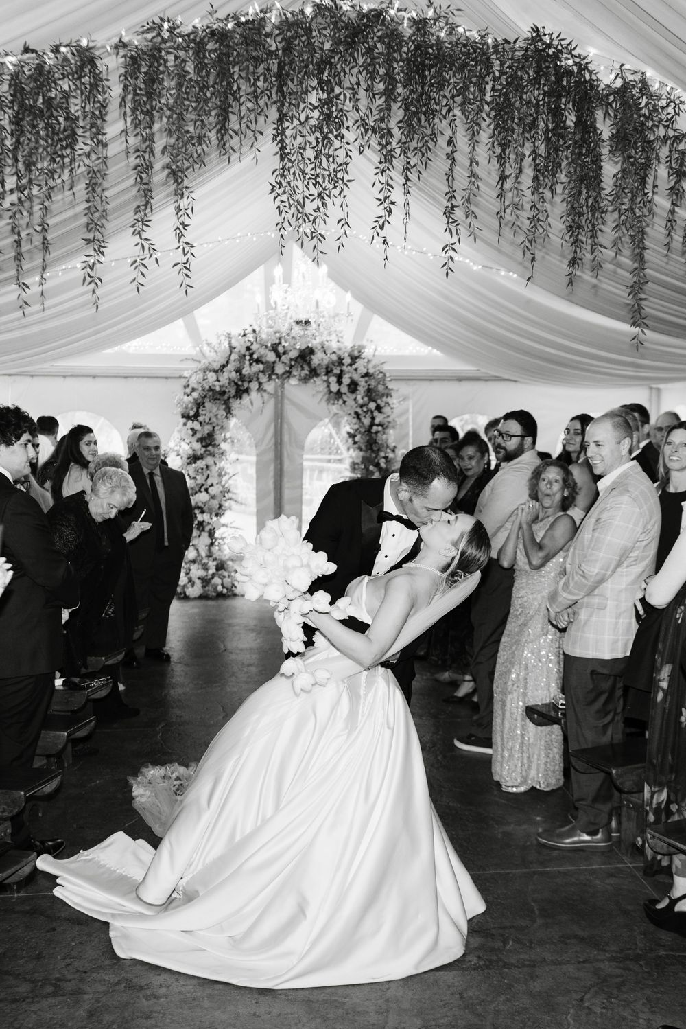 Groom dips bride for romantic kiss after ceremony in black and white wedding photo under floral arch.