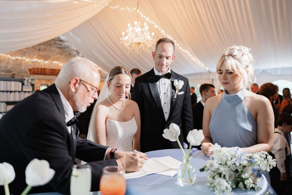 Bride and groom signing wedding documents with officiant and bridesmaid at elegant tented reception.