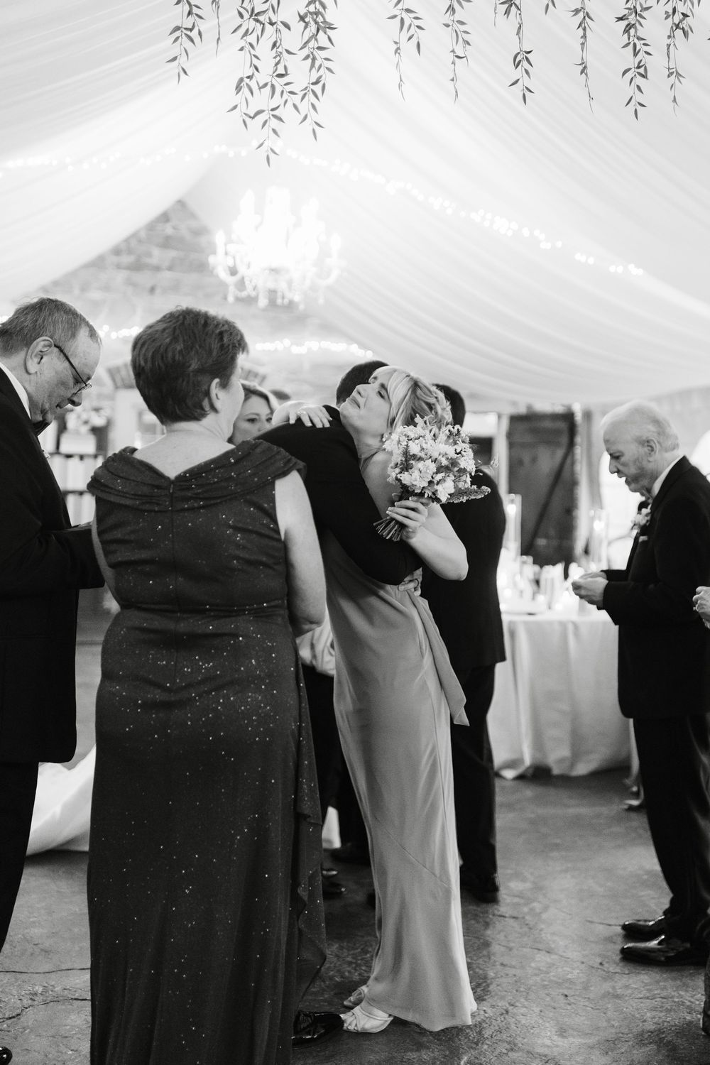 Black and white photo of newlywed bride embracing family member after wedding ceremony in elegant tent.