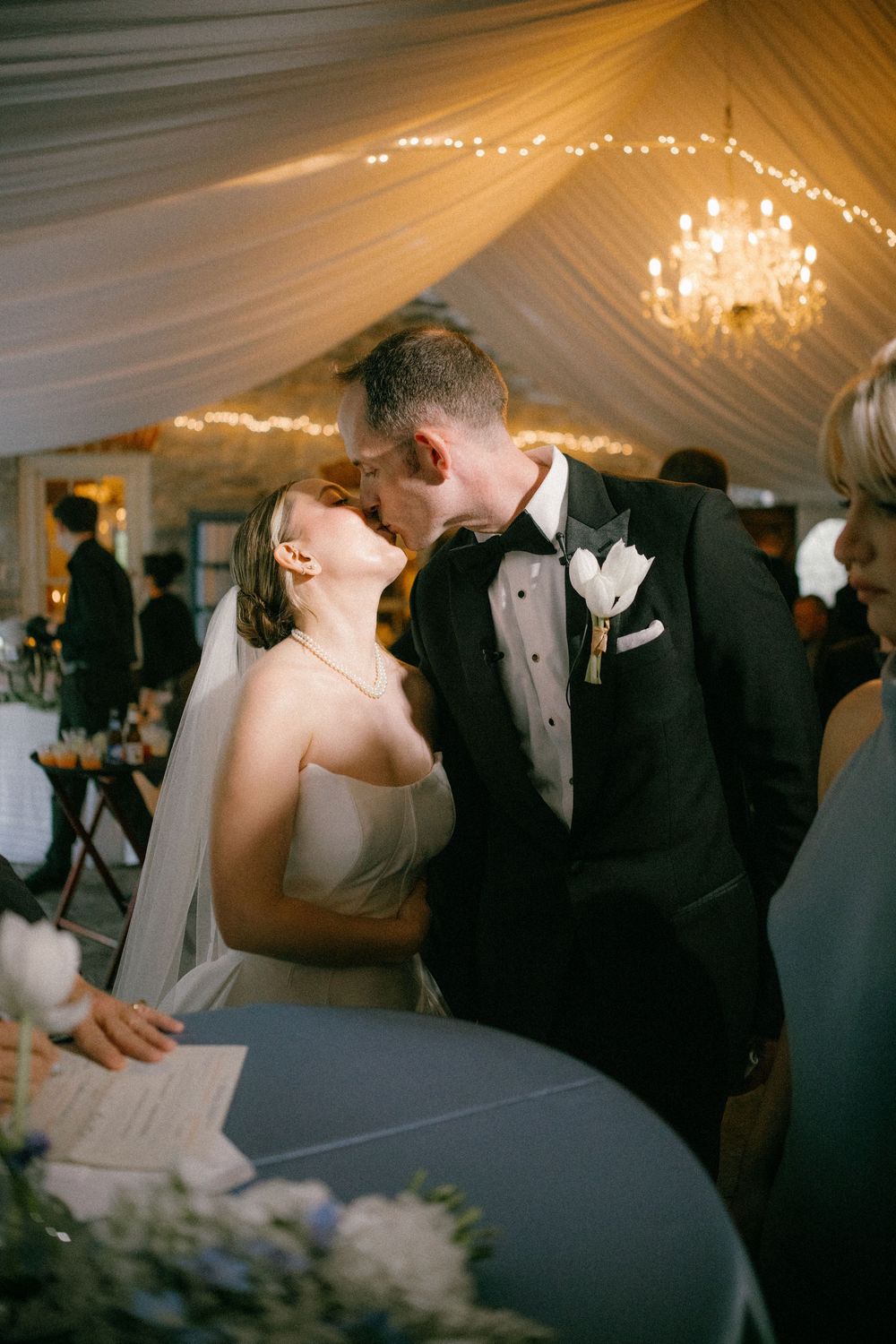 Bride and groom share a kiss at signing table under glowing chandelier in draped reception tent.