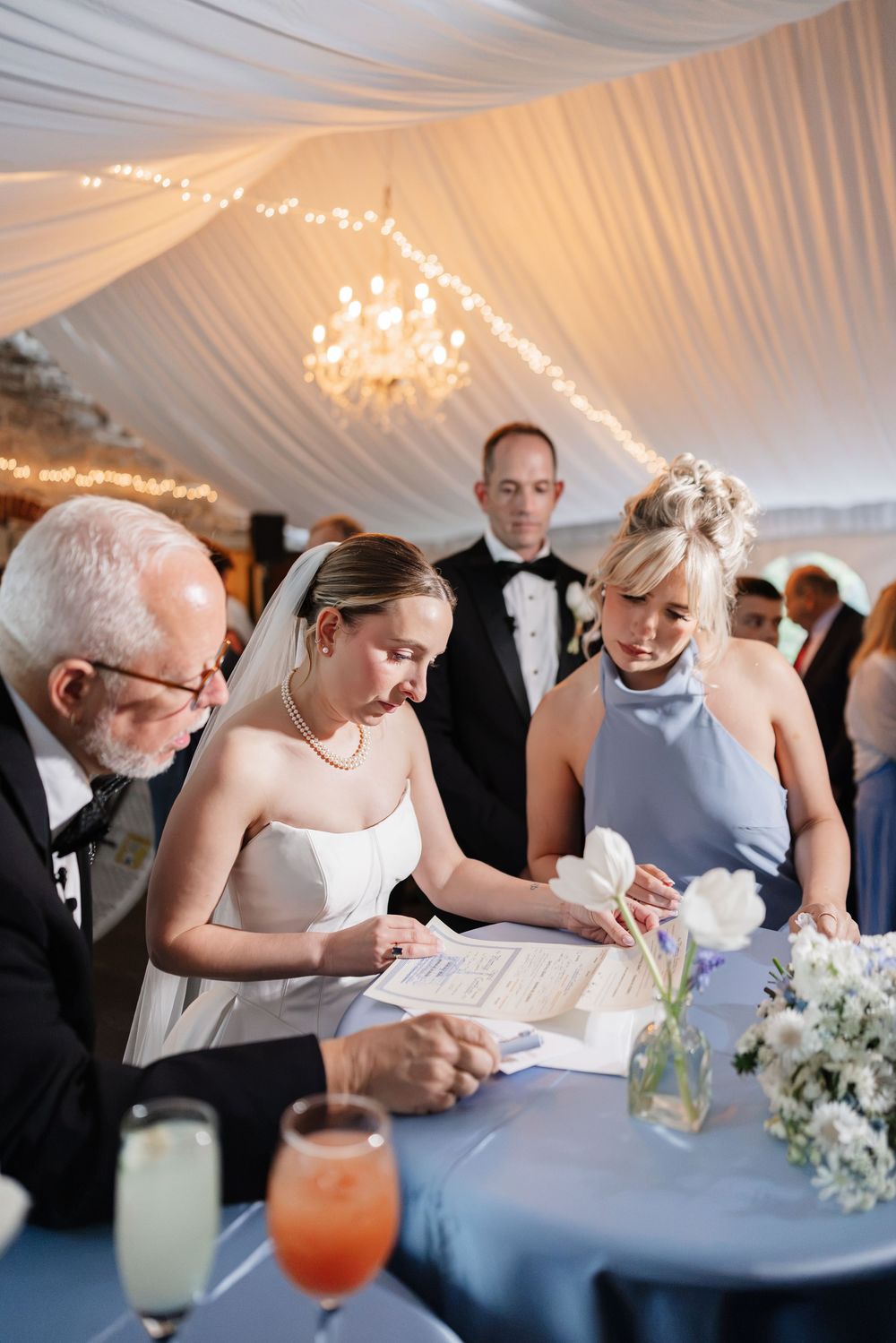 Bride signs marriage certificate at blue-draped table with bridesmaid and guests in elegant tent venue.