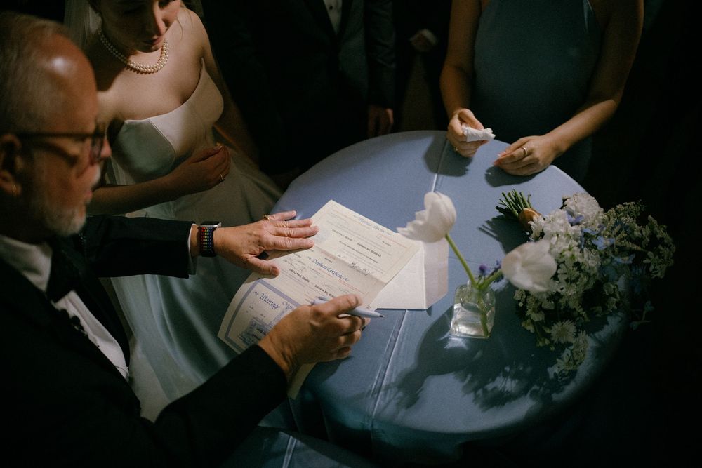 Wedding officiant signs marriage certificate at blue-draped table with white floral centerpiece.
