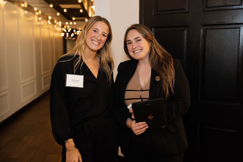 Two women in black attire smile together at an elegant indoor event with string lights in the background.
