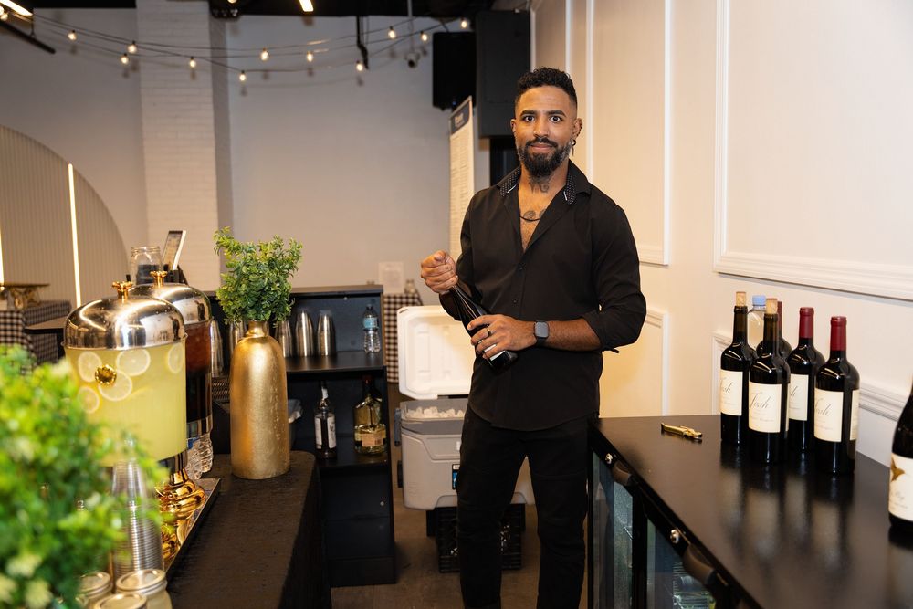 Man in black outfit standing at a bar counter with wine bottles and cocktail equipment in a modern venue.