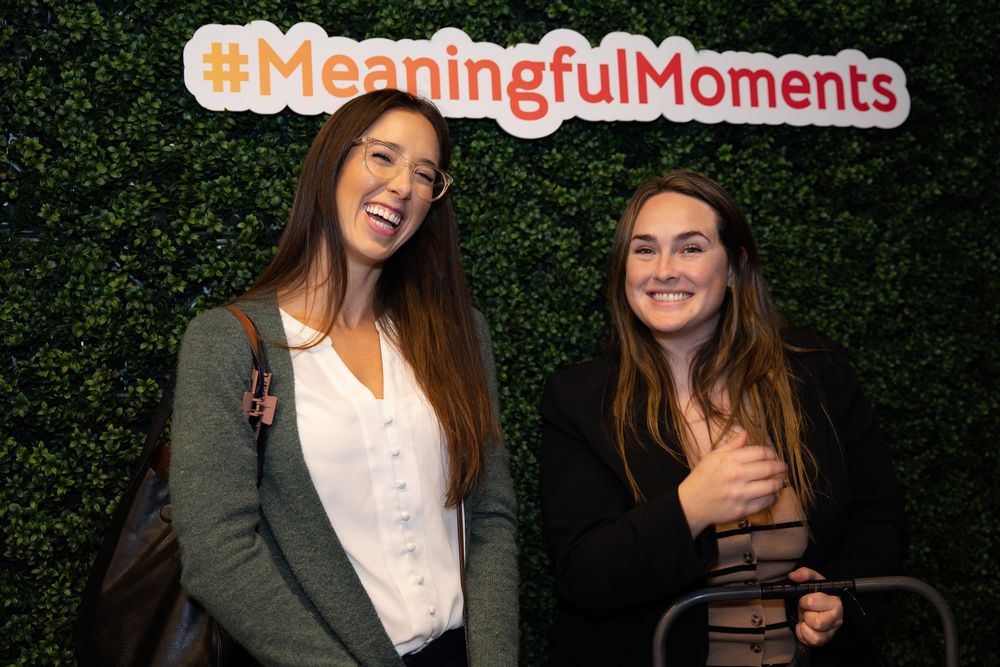 Two women smiling in front of a green hedge wall with a #MeaningfulMoments sign at an event.