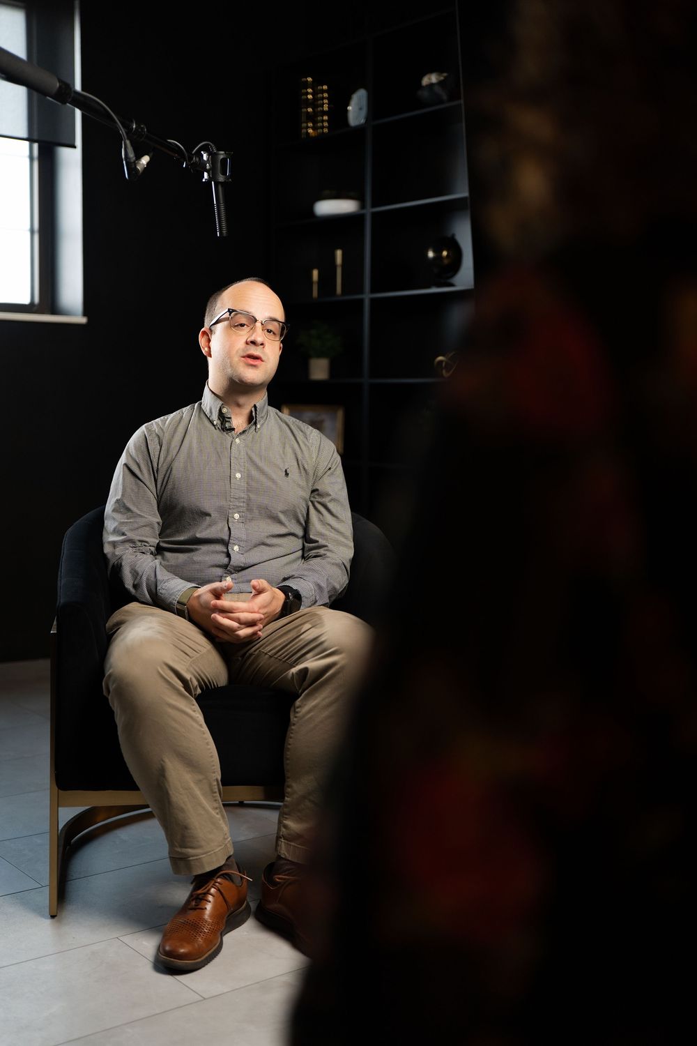 Bald man with glasses sits in black chair, hands clasped, wearing gray shirt and khaki pants in dark office.