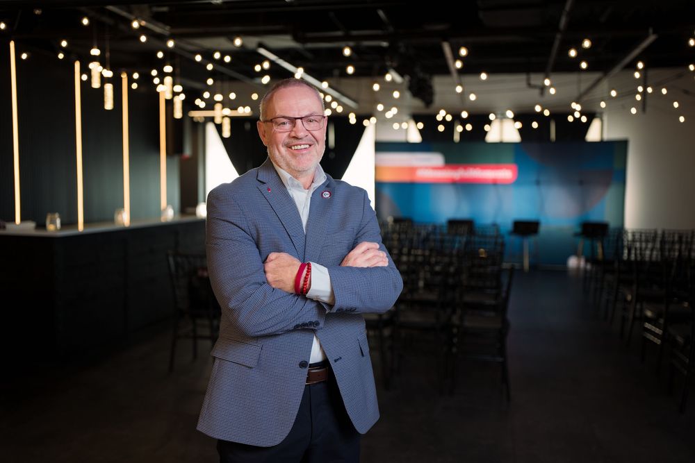 Smiling man in gray blazer stands with arms crossed in modern event venue with string lights.