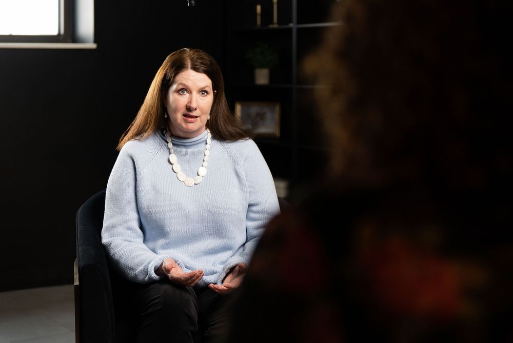 Woman in light blue sweater and pearl necklace seated in a modern dark office setting during an interview.