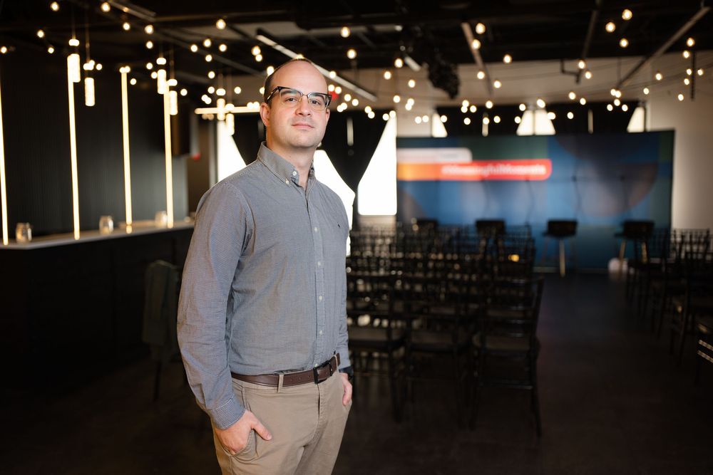 Man in gray shirt stands in modern event venue with string lights and rows of chairs in background.