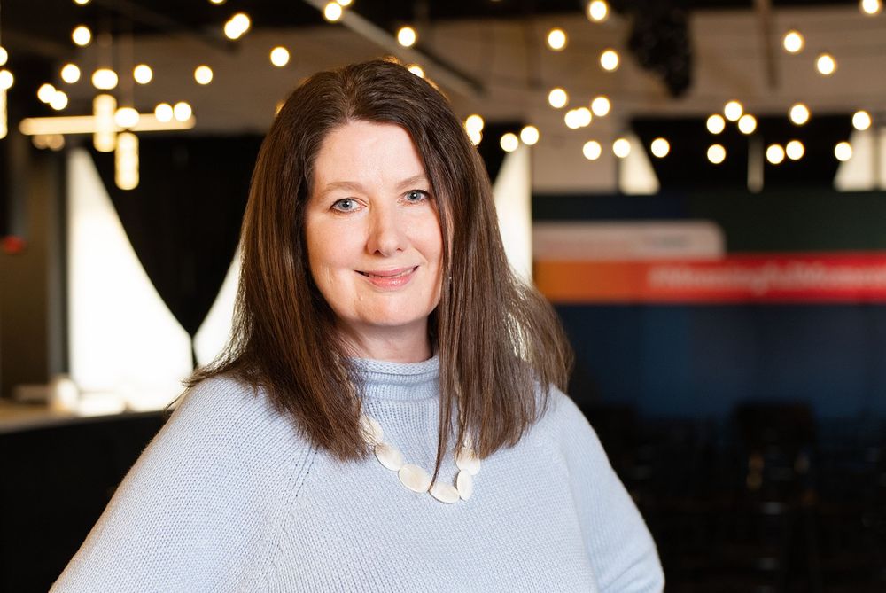 Professional woman with brown hair smiling warmly in a modern venue with decorative string lights.