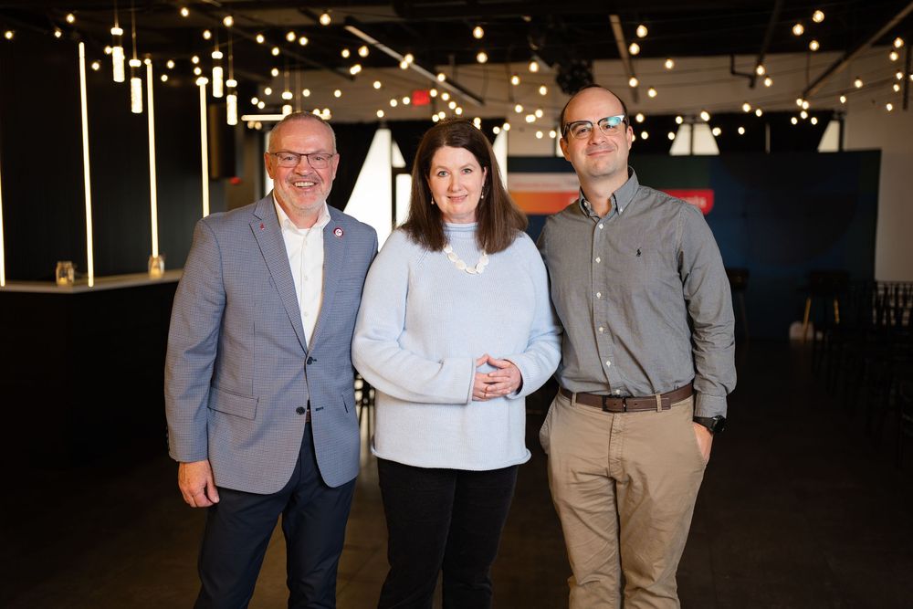 Three professionals smiling and posing together in a modern event space with ambient string lights.
