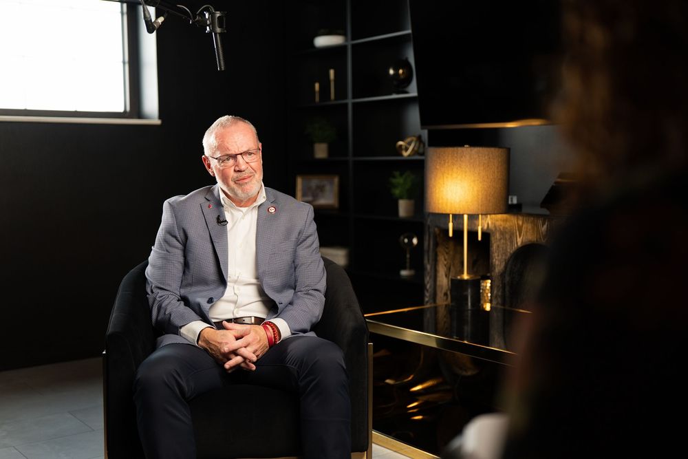 Man in gray suit sitting in black chair during professional interview in modern dark office setting.