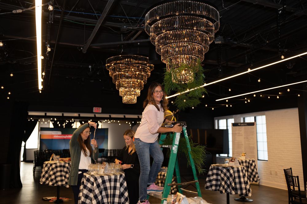 Woman on ladder decorating elegant chandelier in event venue with black and white checkered tablecloths.