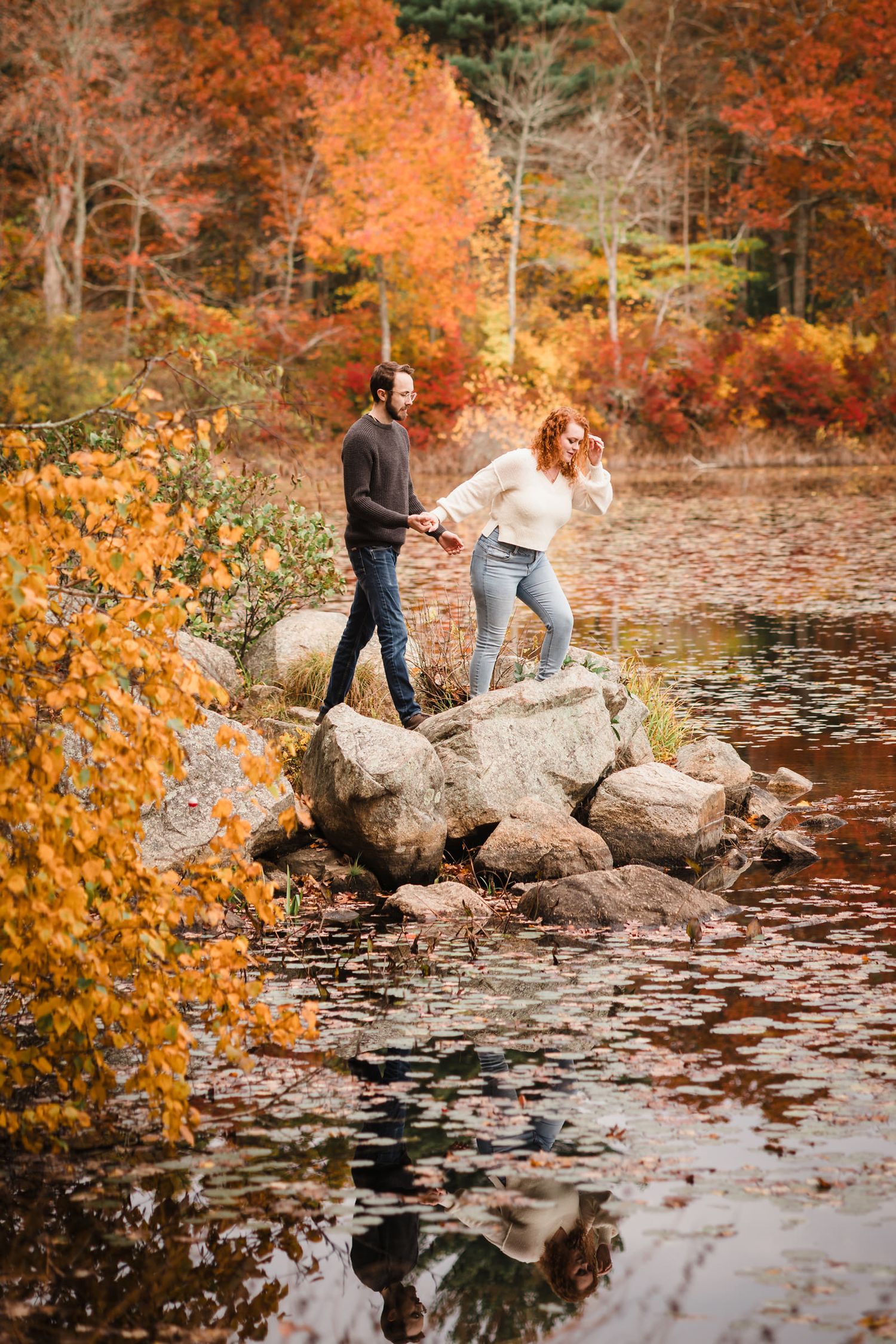 Engaged couple holding hands while carefully crossing rocks at the edge of Lake Leach, surrounded by fall color and water reflections.