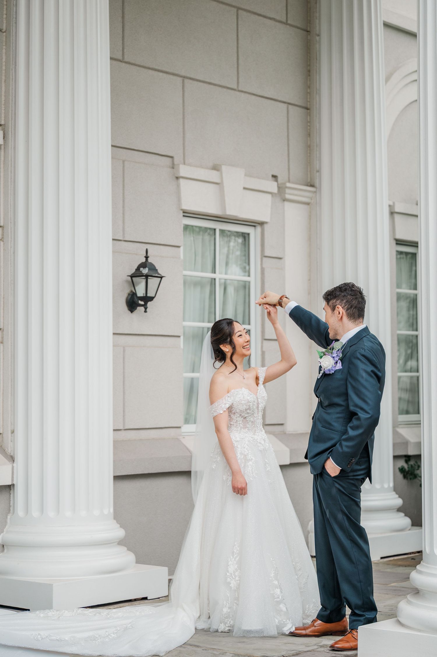 Bride and groom dancing on elegant white columned venue steps, bride in lace off-shoulder gown, groom in navy suit.