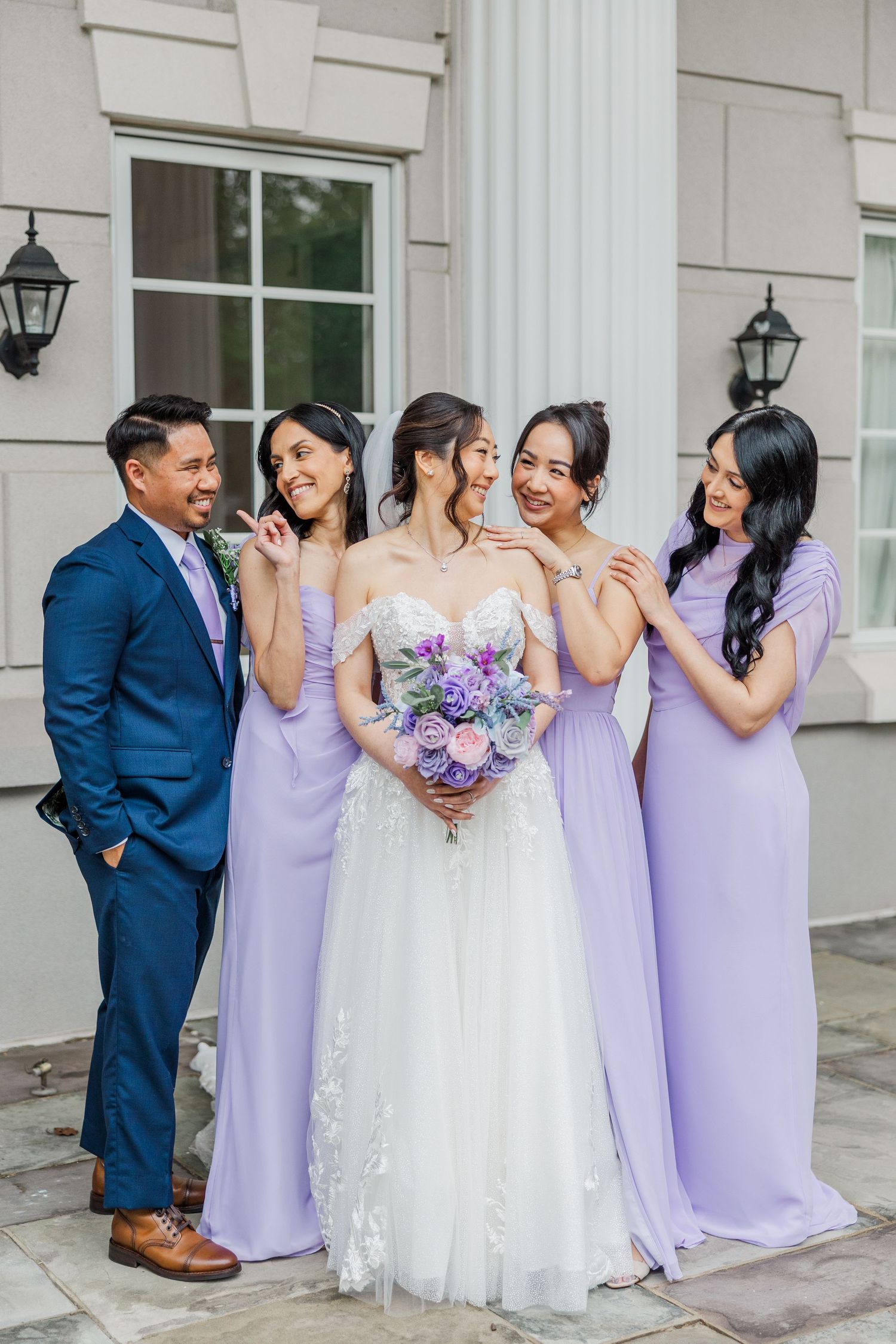 Bride in white lace gown holds purple bouquet surrounded by bridesmaids in lavender dresses and a groomsman in navy suit.