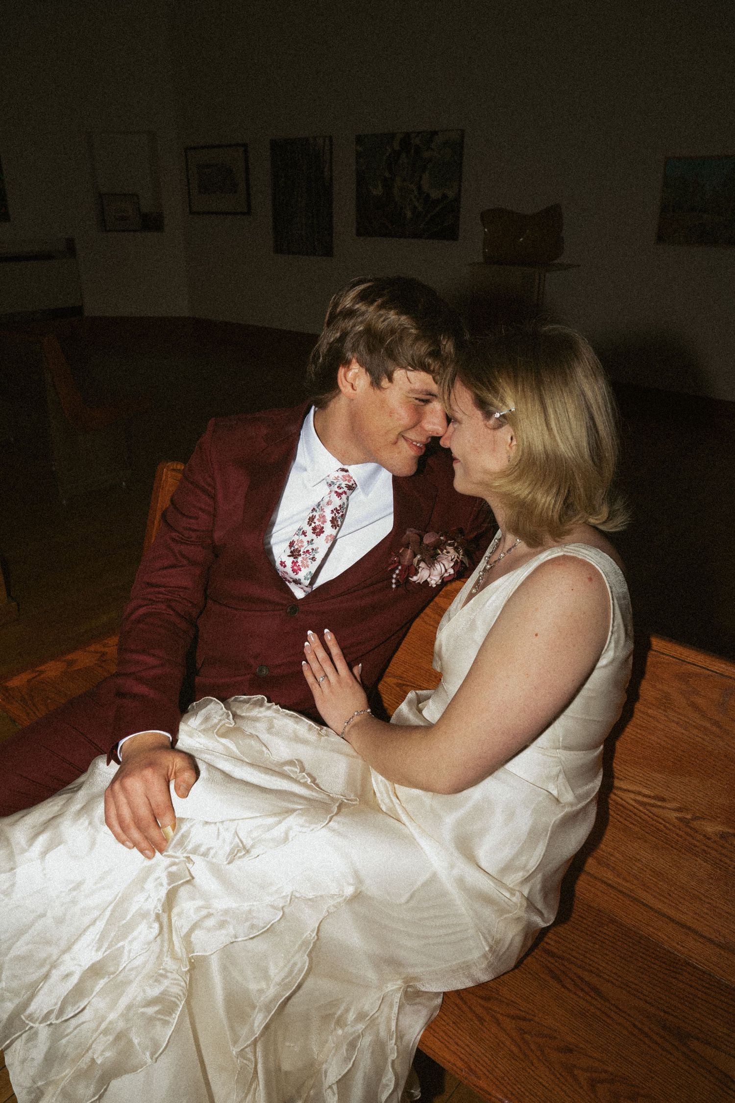 Bride and groom in wedding attire share an intimate moment, sitting close together in a warmly lit room.