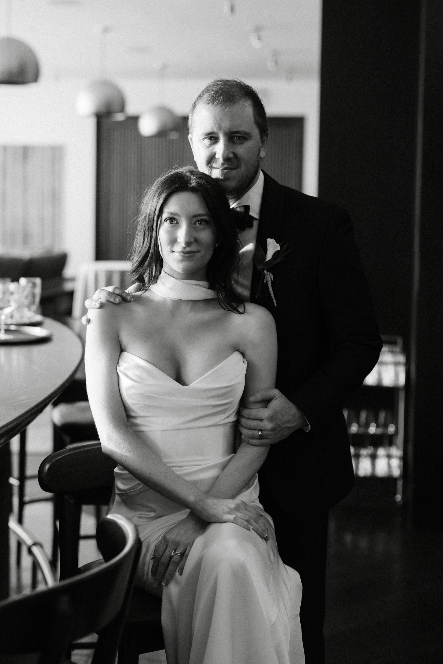 Black and white wedding portrait of bride in strapless gown seated while groom in suit stands behind her.