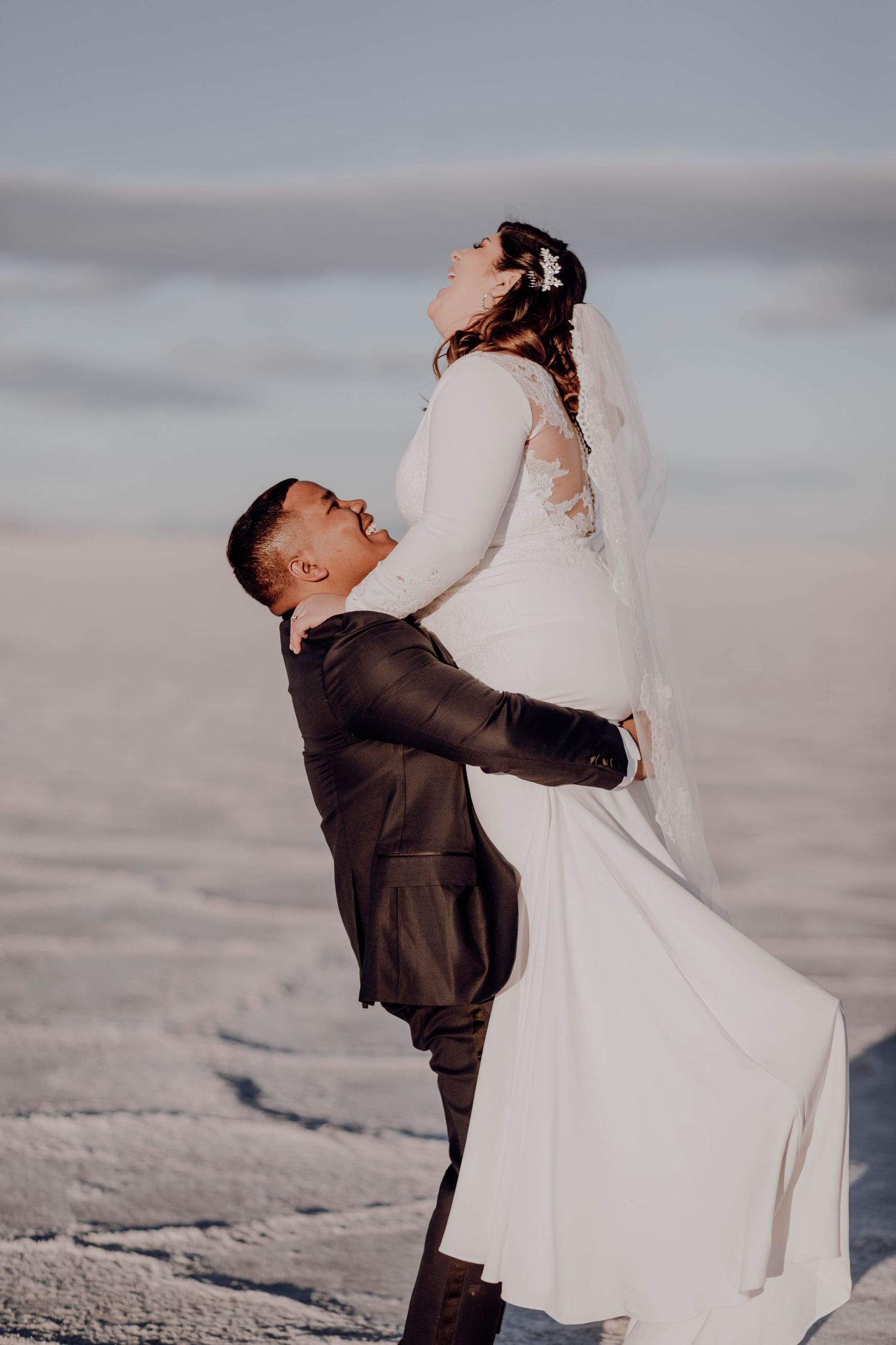 Groom joyfully lifting bride in white gown on snowy salt flats under a clear blue sky.