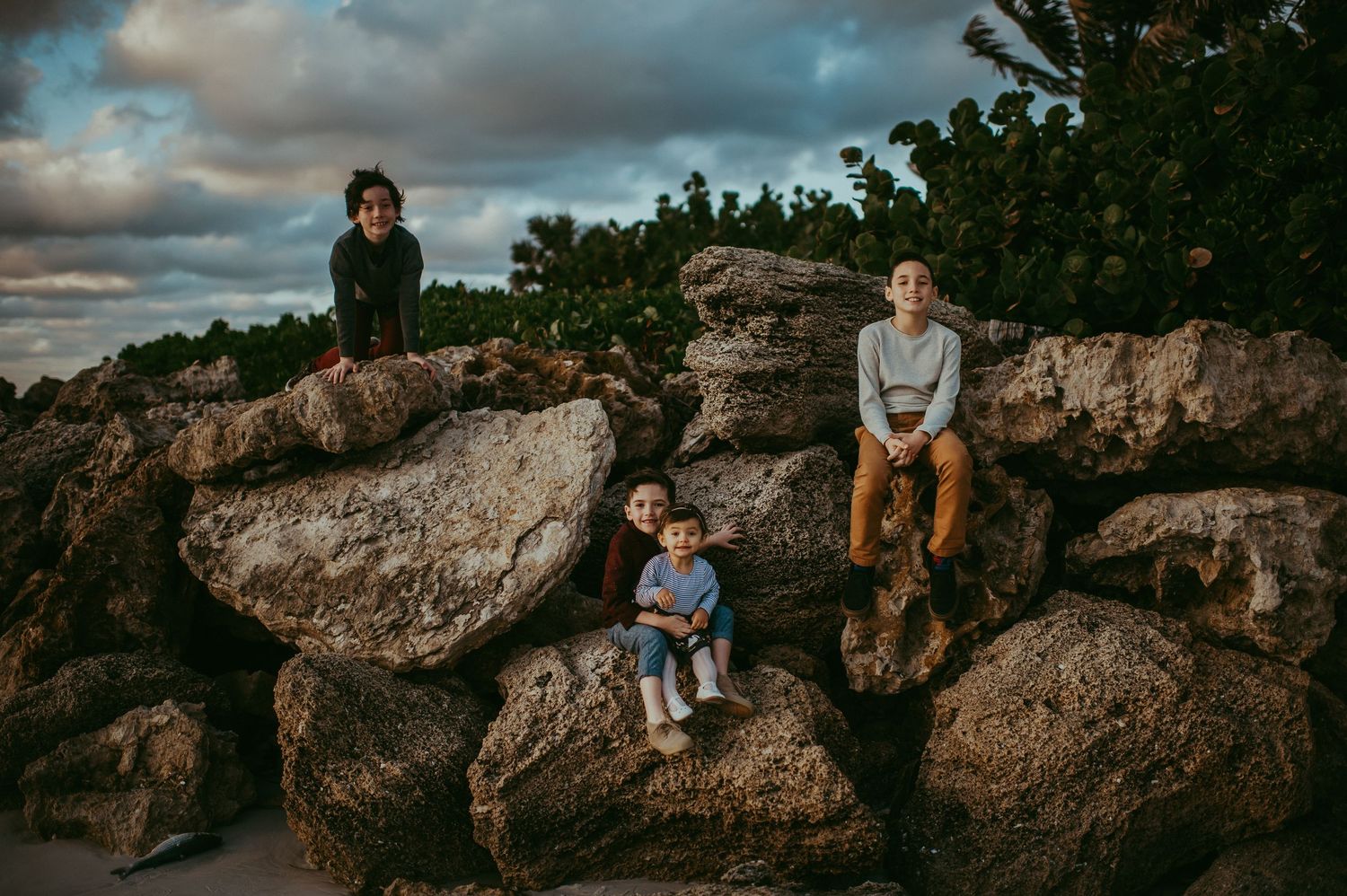 Four children sitting and climbing on large rocks outdoors under a cloudy sky near trees.