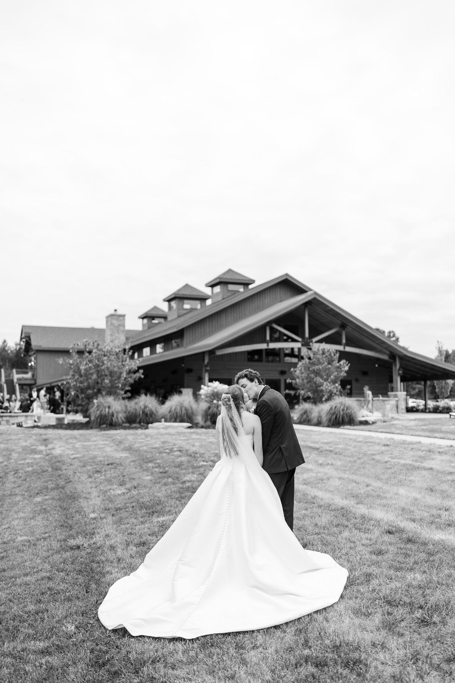 Black and white photo of bride and groom kissing on lawn before rustic wedding venue.