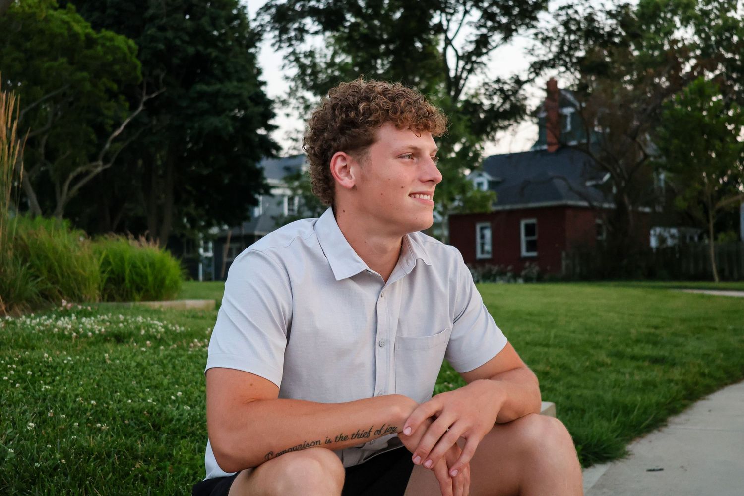 Young man with curly hair sitting on grass in park, smiling outdoors near brick building on sunny day.