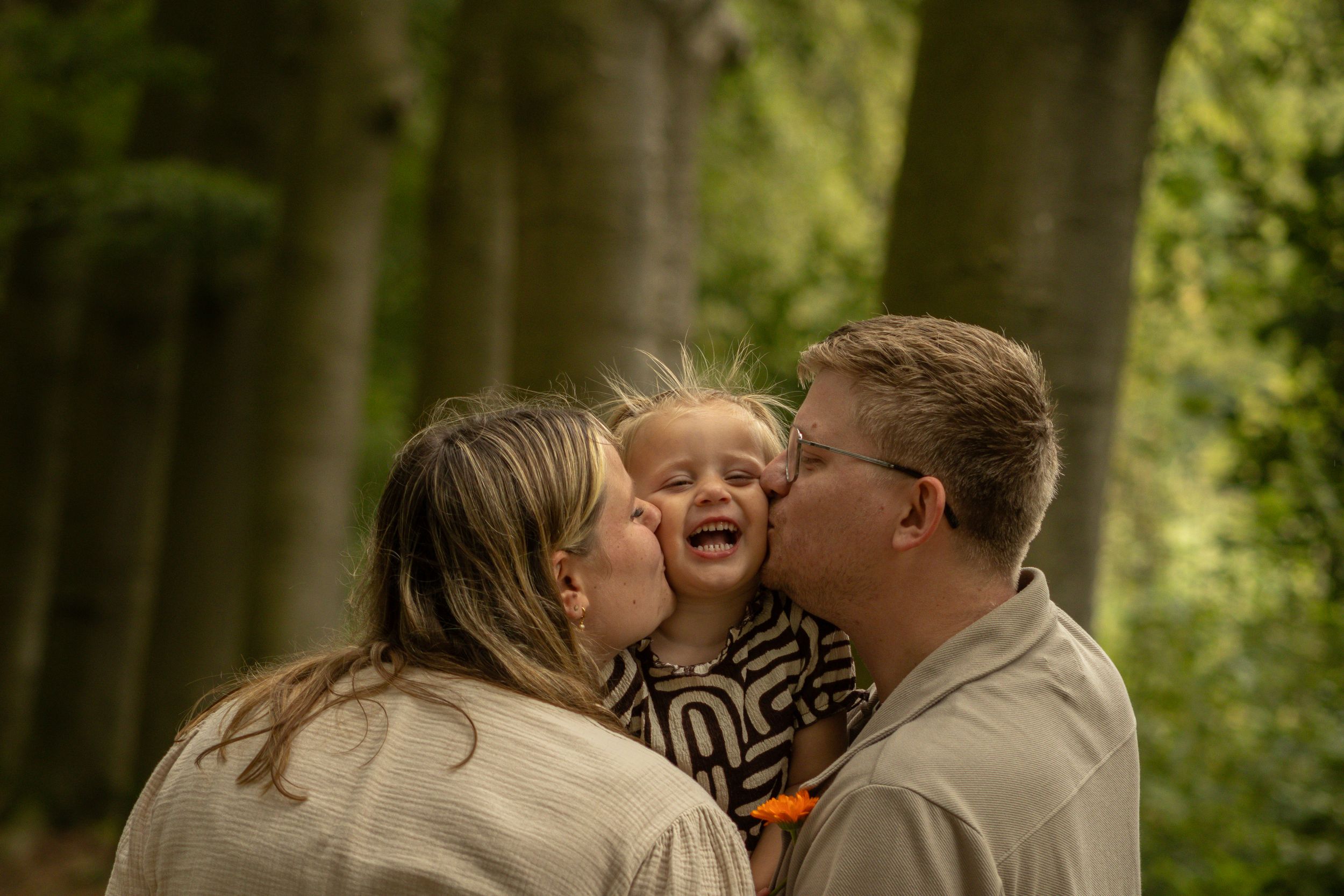 Twee ouders geven hun lachende kind tegelijk een kus op de wangen in het bos.