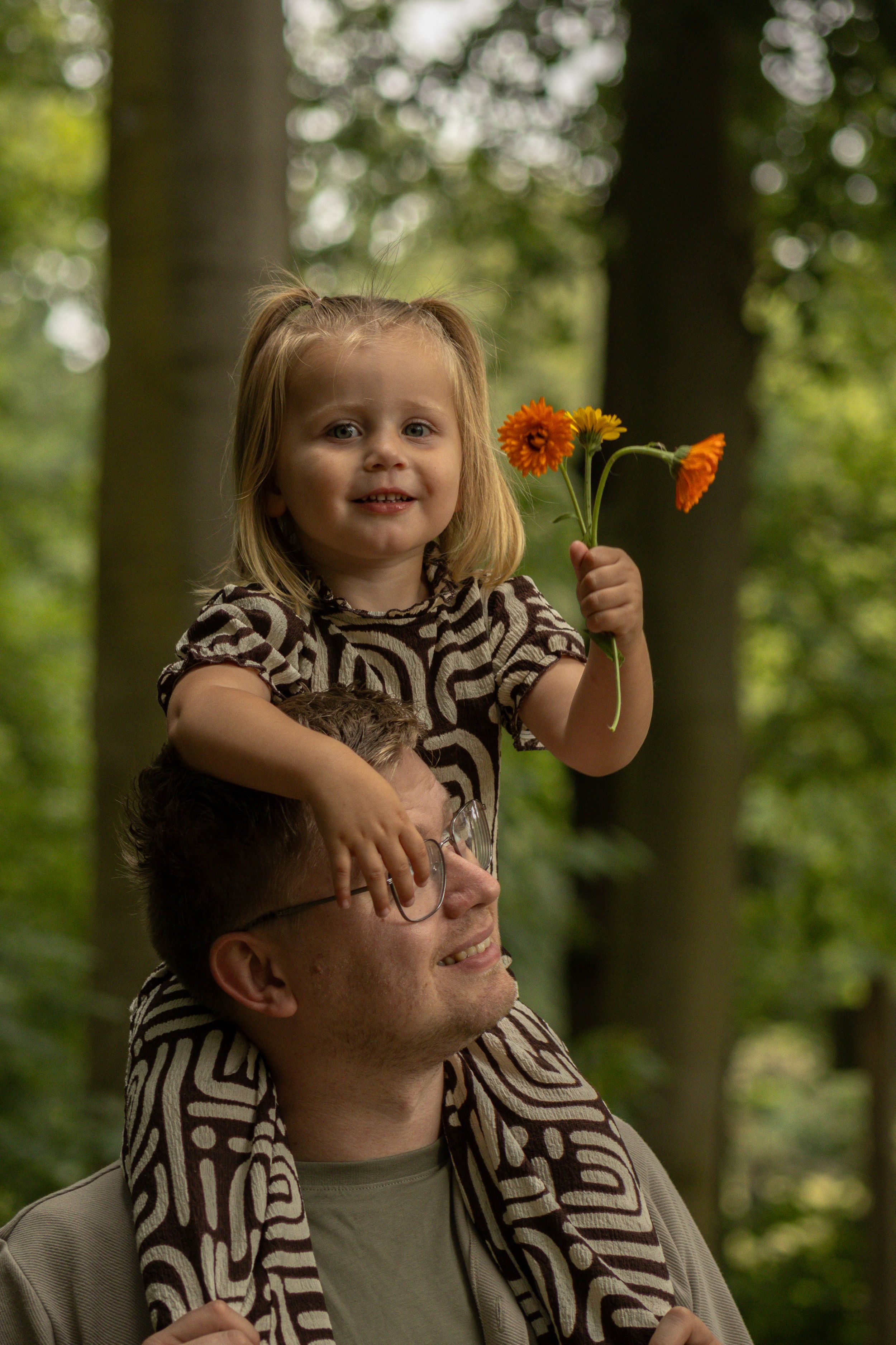 Lachend kind zit op de schouders van een ouder en houdt twee oranje bloemen vast in het bos