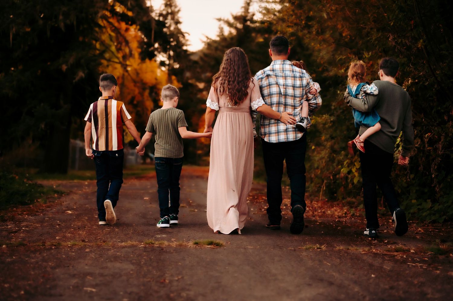 Family of six walking hand-in-hand down a scenic autumn path surrounded by colorful fall foliage.