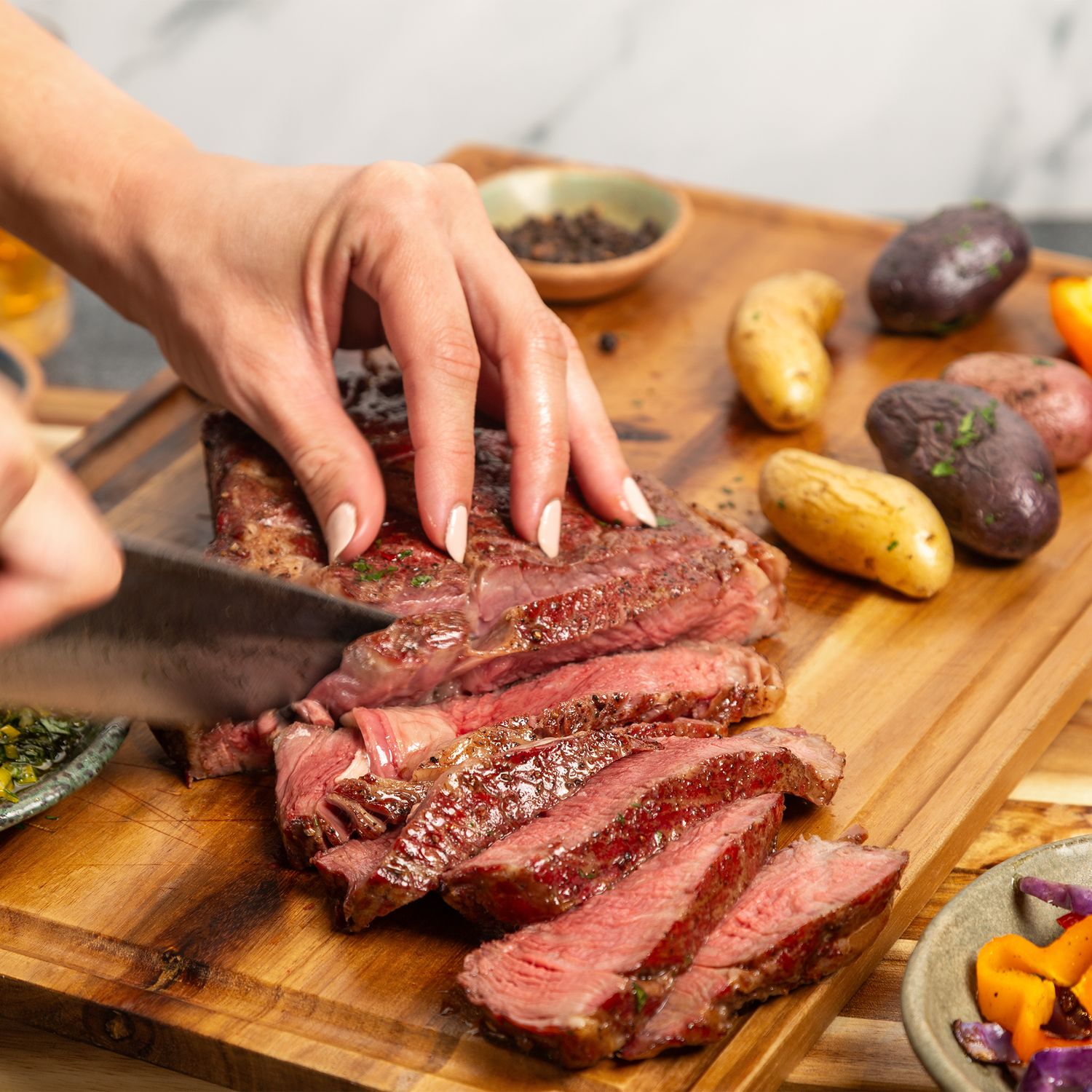 Person slicing medium-rare grilled steak on wooden cutting board with colorful roasted vegetables.