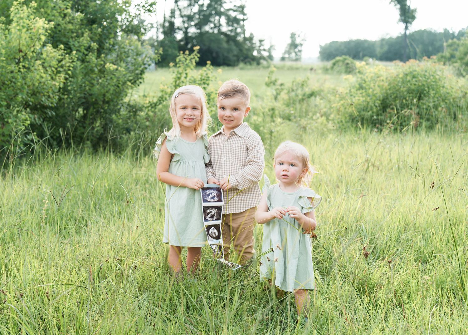 Three young siblings pose in a lush green meadow, dressed in coordinating mint and tan outfits for a family portrait.