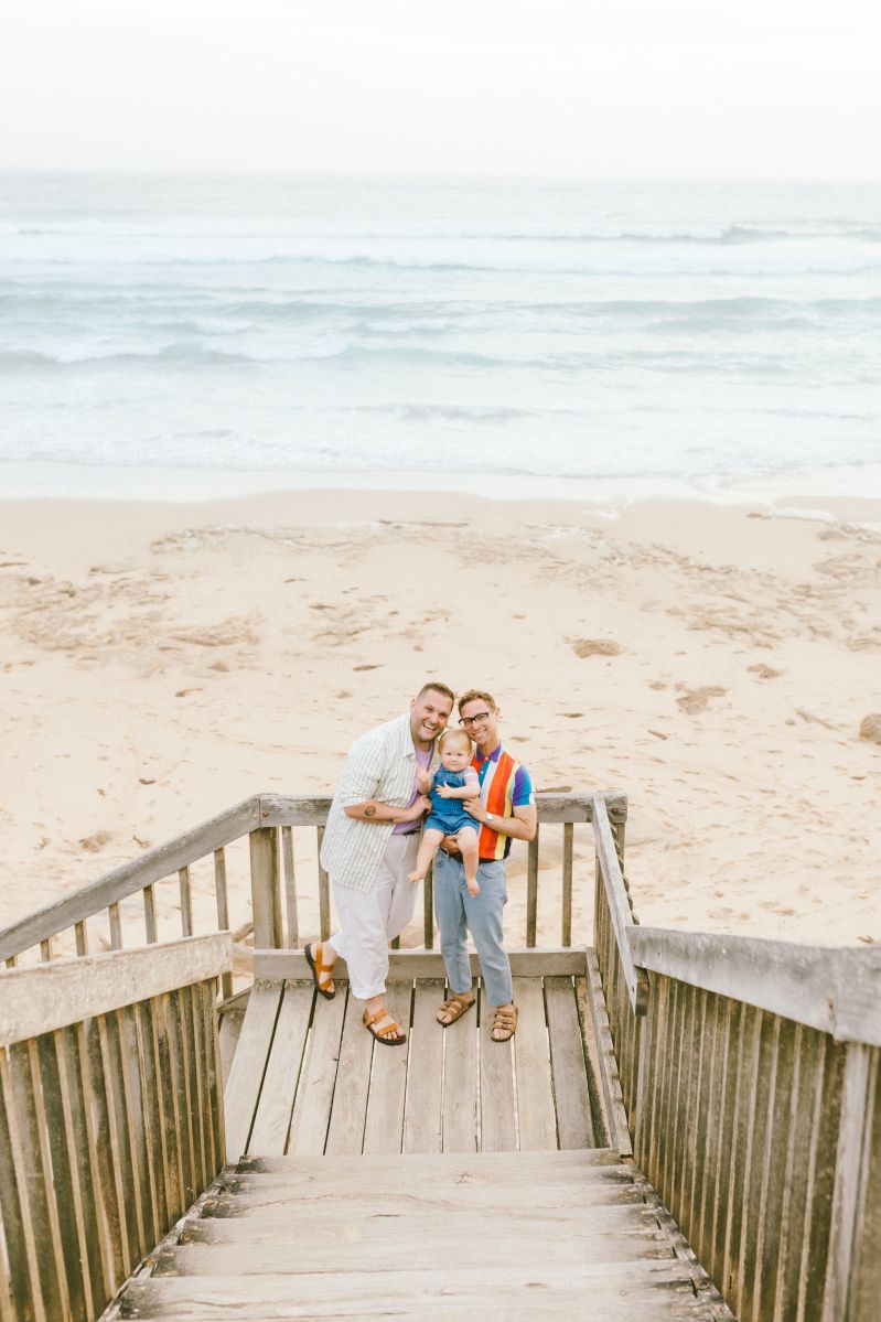 Two men holding a baby on a wooden beach boardwalk with sandy shore and ocean waves in the background.