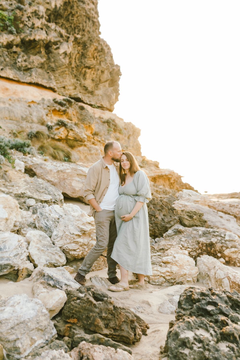 A couple poses on rocky coastal cliffs, the man kissing his pregnant partner's head as she cradles her baby bump.