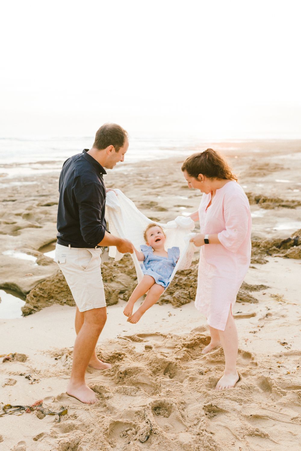 Family of three playing on sandy beach at sunset, parents swinging laughing toddler in white blanket near rocky shoreline.