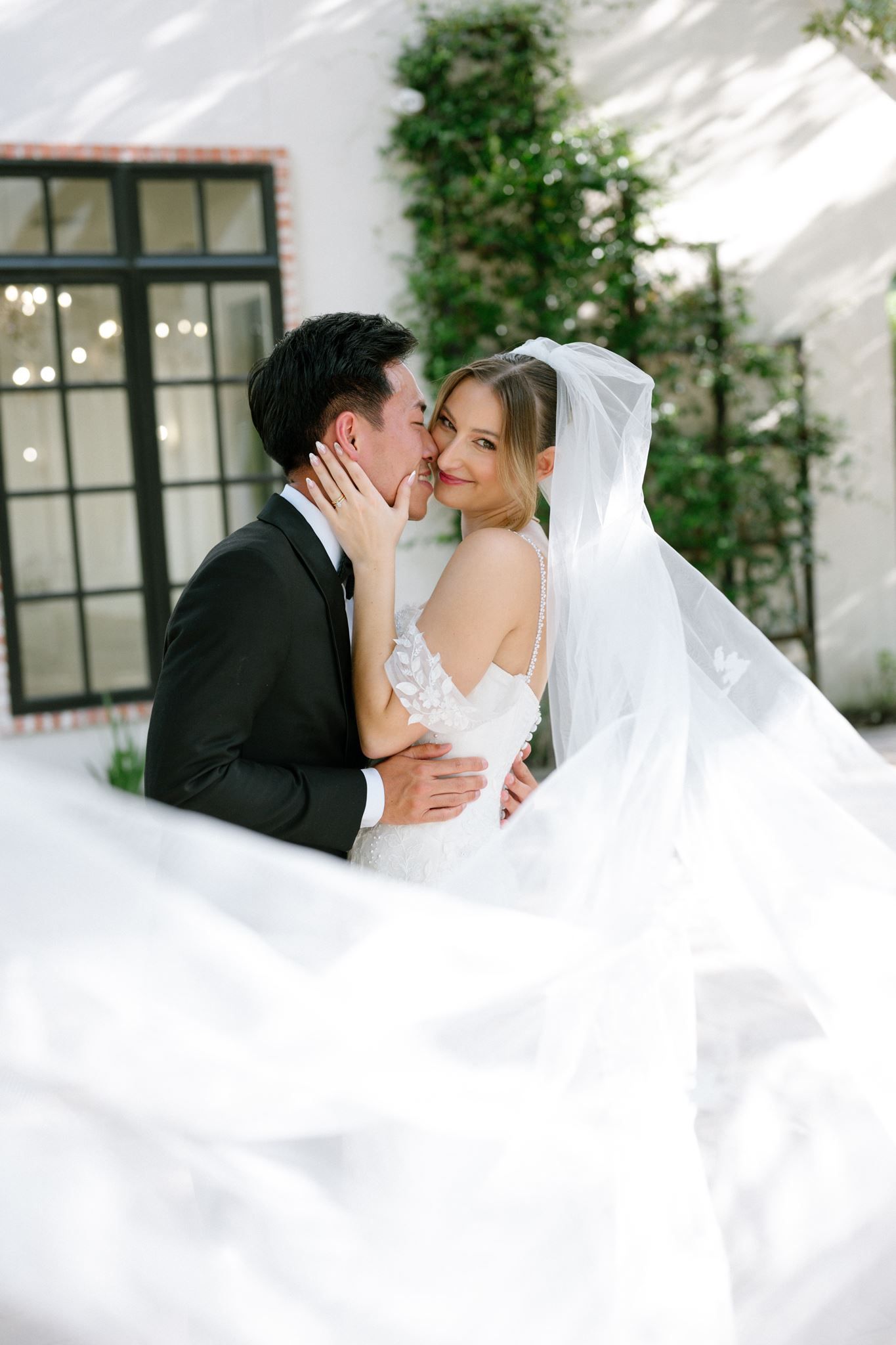 Bride and groom share a romantic kiss outdoors, with her flowing white veil billowing around them near an ivy-covered wall.