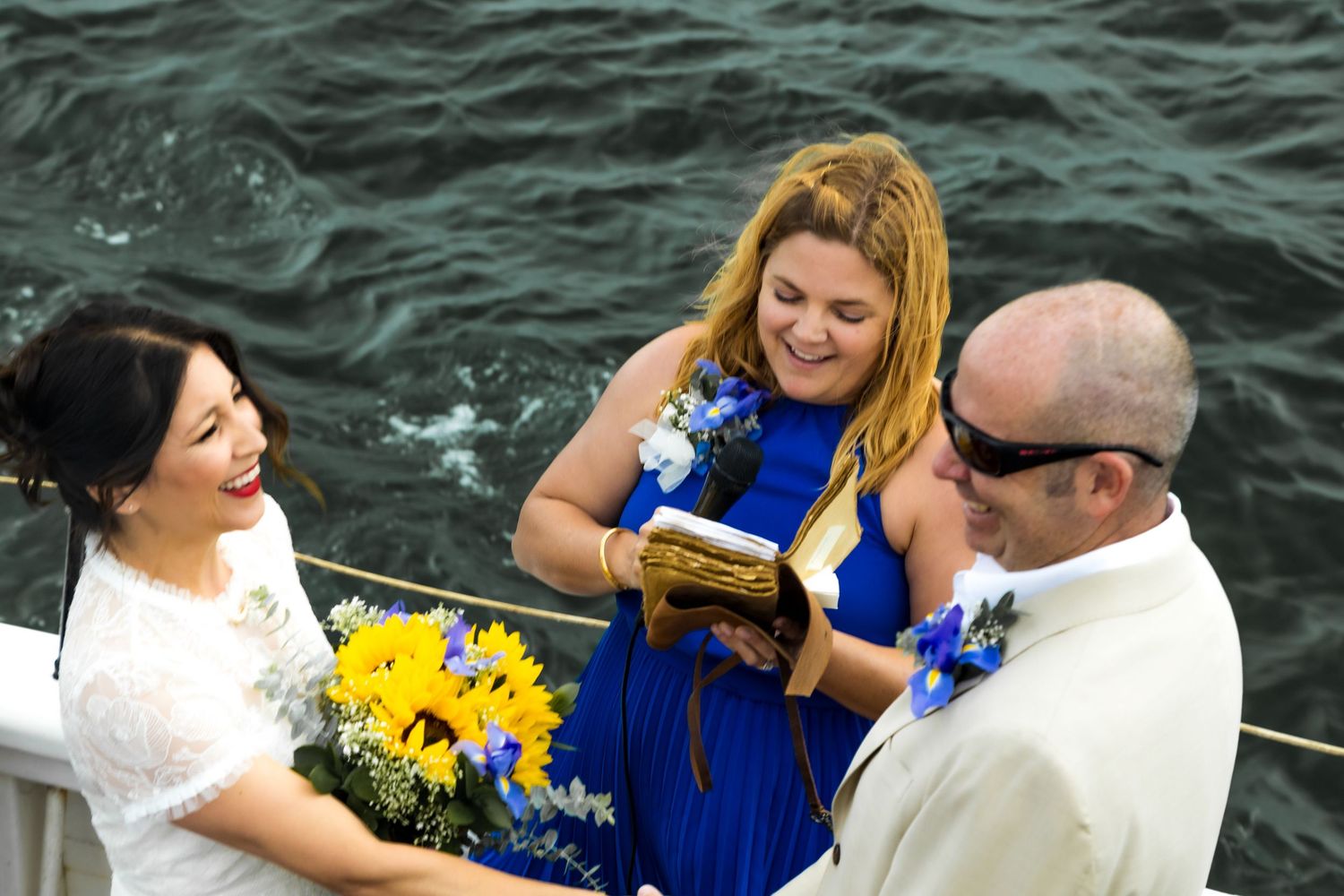 Outdoor boat wedding ceremony with bride holding sunflowers, groom in blue boutonniere, and officiant in blue dress reading vows.