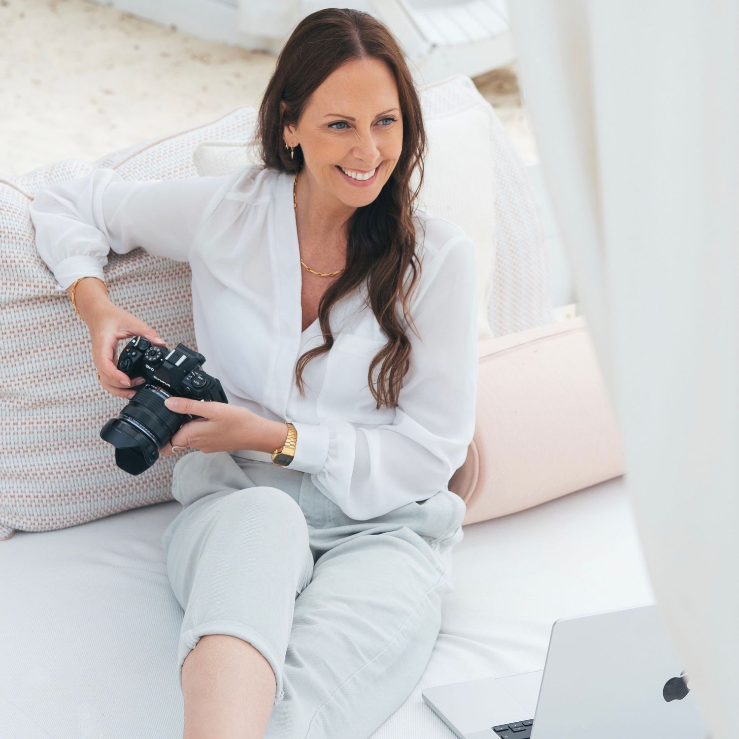 Smiling woman in white shirt sitting on floor holding a DSLR camera in a bright, airy room.
