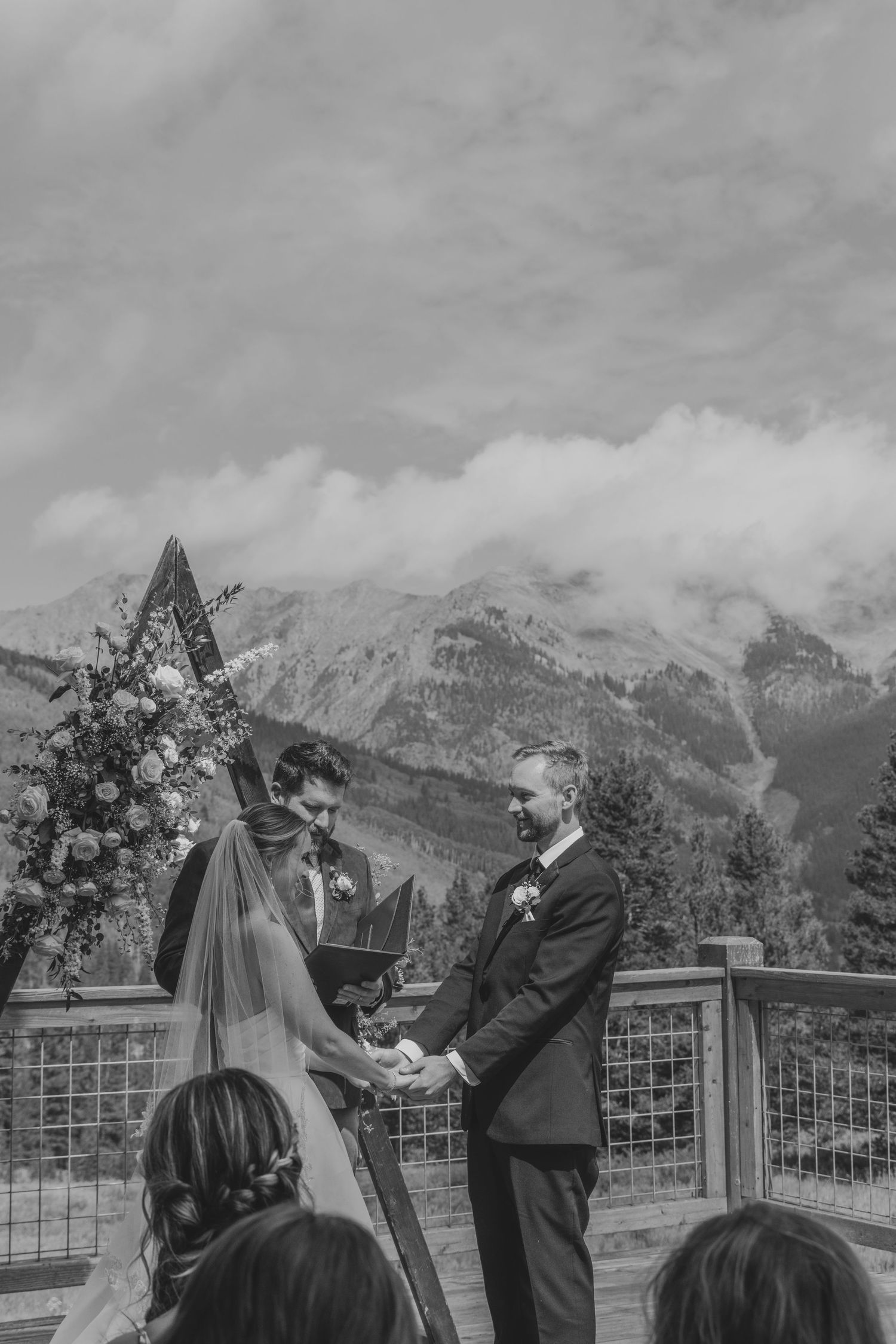 Black and white outdoor mountain wedding ceremony with couple exchanging vows at floral arch with snowy peaks backdrop.