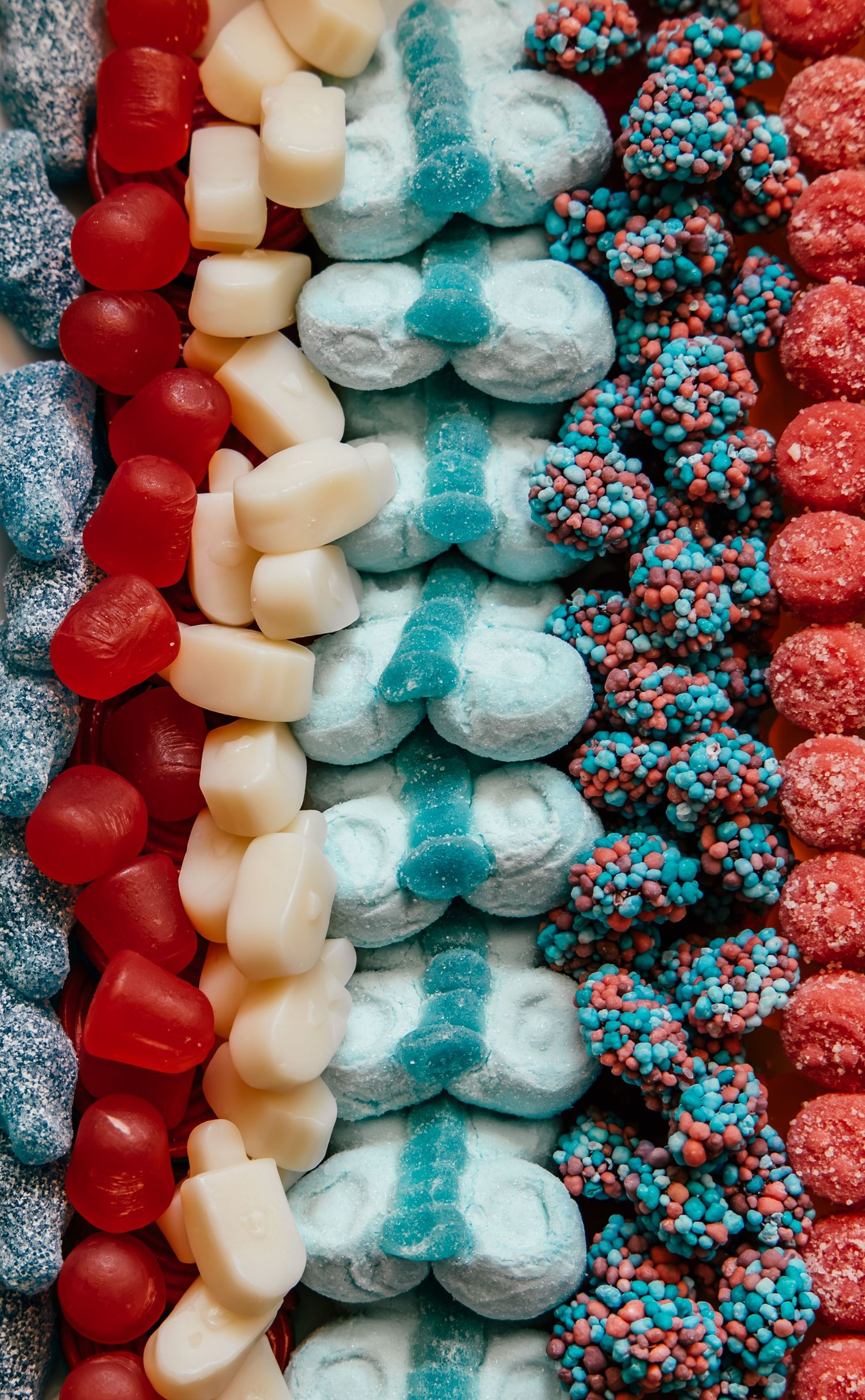 Colorful rows of assorted candies including red gummies, white marshmallows, blue sugar bears, and sprinkled chocolate clusters.