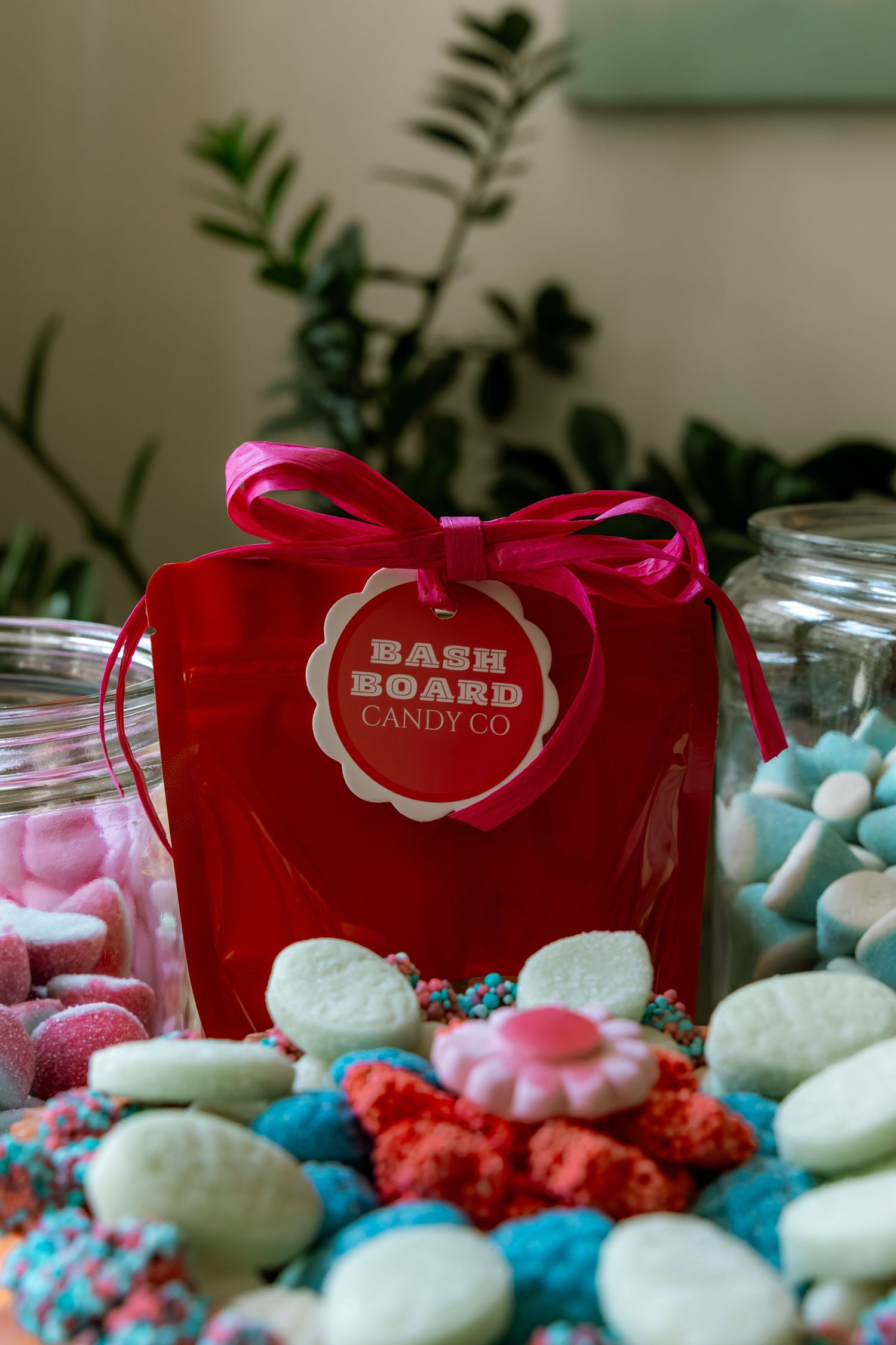 Red Bash Board Candy Co gift bag with pink bow surrounded by colorful assorted candies and glass jars on a table.